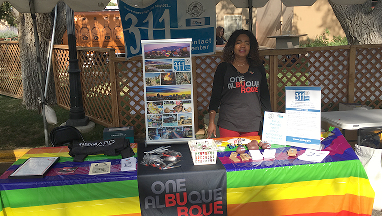 A 311 booth with various posters and print material with the 311 and OneAlbuquerque logo, with a woman wearing a One Albuquerque shirt smiling at the camera.