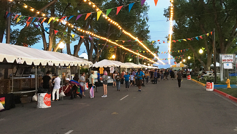 A group of people at the state fair, browsing shops along a concrete road lined with trees and decorated with strands of lights and colorful flags.