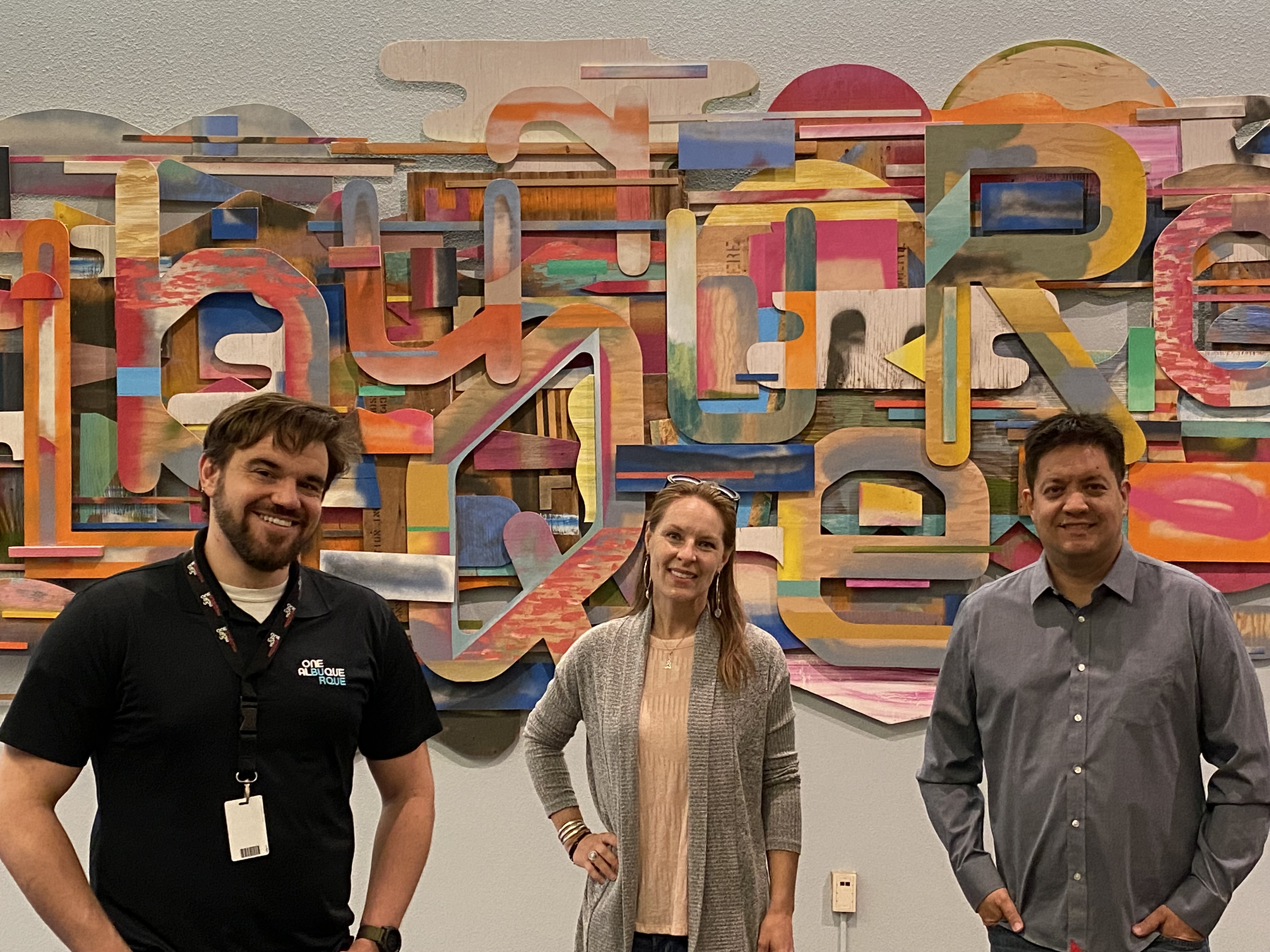 3 Office of Civic Engagement employees facing towards the viewer standing in front of a colorful cloud with the word Albuquerque spelled out within it. From left to right: Nicholas Vottero, Mariah Harrison, and David Chene