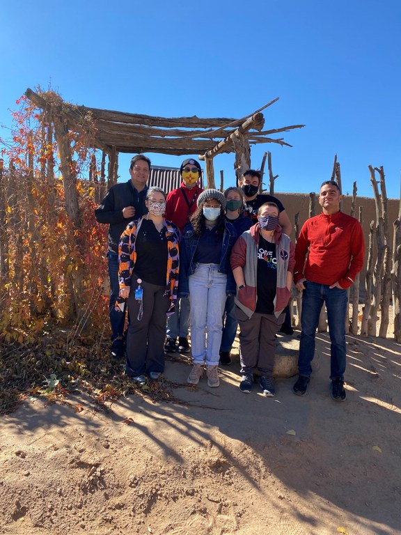 8 people facing the viewer, posing as a group in front of a stick fence and gate. 4 people stand in the front row with their hands in their pockets, 4 people stand behind them, with faces visible.