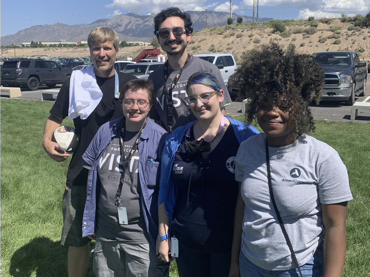 5 people posing for a photo, facing the viewer. 2 people in the back, on the left side of the image with 3 individuals in front, on the right side of the image. Mountains, a hill, a parking lot, and grass are in the background.