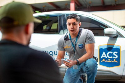 Male ACS Responder kneeling in the background with an ACS vehicle behind him, talking to a man whose back is out of focus in the foreground.