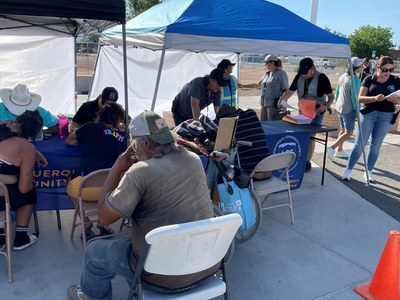 A bustling Connect-to-Care pop-up event with unsheltered individuals seated at tables under tents, engaging with Albuquerque Community Safety (ACS) team members. ACS staff in uniforms assist participants with paperwork and provide resources, while other staff stand nearby, coordinating activities. The setup includes informational materials on tables and a visible ACS banner, emphasizing outreach and community engagement.