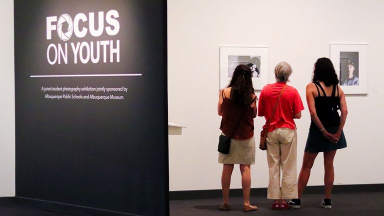 Three women looking at a picture hanging on the wall next to a sign that says "Focus on Youth a juried student photography exhibition jointly sponsored by Albuquerque Public Schools and Albuquerque Museum.