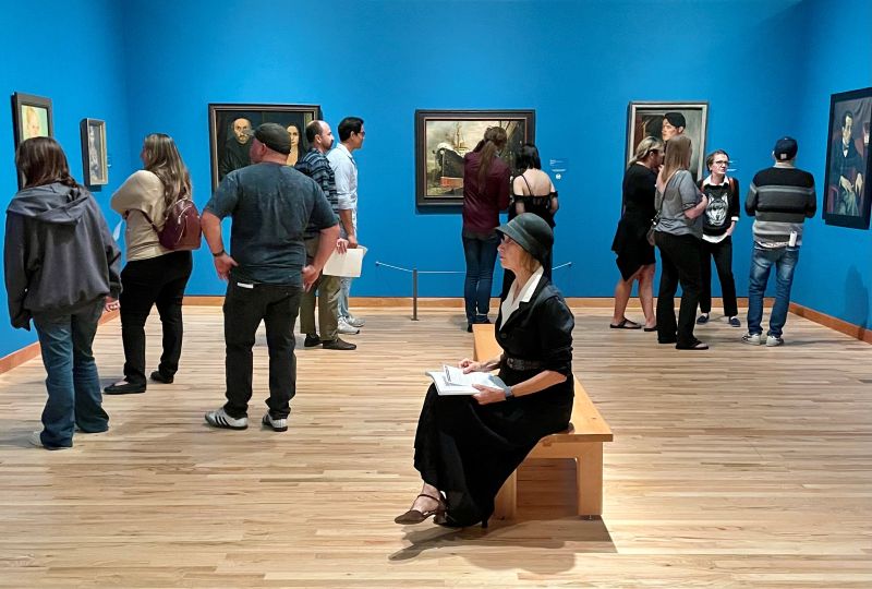 Museum gallery image of a space painted gray with a central bronze scuplture of a bronze male nude on a rectangular pedestal surrounded by viewers pointing at it and talking with one another.