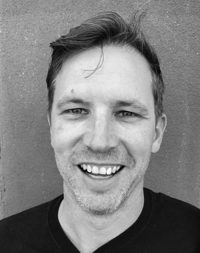Karl Hofmann Headshot A black-and-white headshot of artist Karl Hofmann, a man with light-colored skin, short, dark hair, and stubble, pictured smiling in front of a grey background.