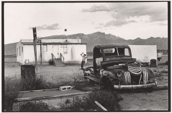 Danny Lyon Llanito A vintage black and white photo of a boy playing basket ball outside his mobile home. There is a run-down care in the foreground and mountains in the background.