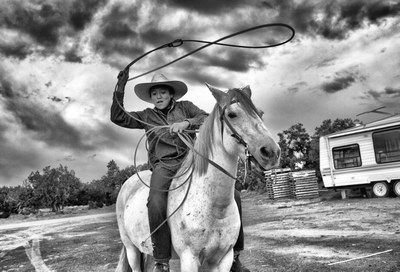 A black-and-white photograph of a young man on horseback, wearing a cowboy hat and swinging a lasso above his head.