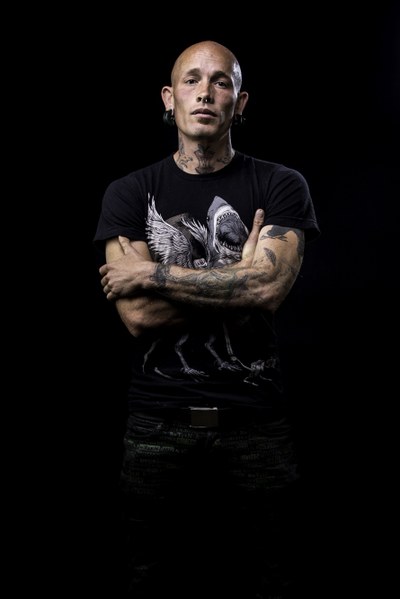 A portrait of artist Ernest Doty standing in front of a black background. Doty looks forward with his arms crossed and is bald light-colored skin and tattoos on his arms and neck. He is wearing a black T-shirt with an image of a shark on it.