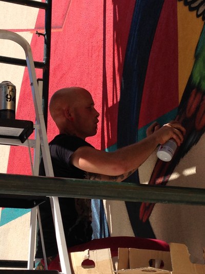 An image of artist Ernest Doty using spray paint to create a mural of a brightly colored bird.