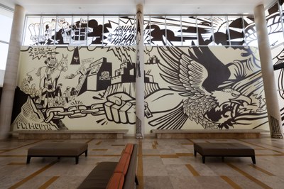 A mural on the wall and window of the Albuquerque Museum lobby. On the left is a soldier on horseback watching a line of people walking into an ice box, beneath a covered wagon, a ship, and a crawling robot asking for water. In the center is laughter coming from pueblo buildings. On the left is the Statue of Liberty in chains, being bit by an eagle.