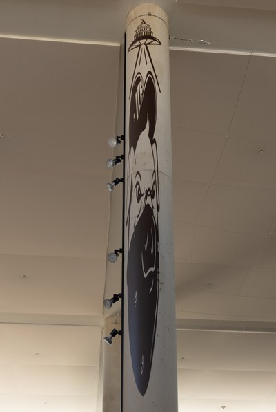 A black-and-white image on a column in the Albuquerque Museum Lobby, depicting a pig wearing a face mask and a hat labeled "ICE" being abducted by a flying saucer with the US Capitol Building in place of a dome, stretched in anamorphic perspective.