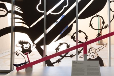 A mural on the window of the Albuquerque Museum lobby. The image depicts two children interacting with Teeter-Totter Wall (2019) by Ronald Rael, Virginia San Fratello, and Colectivo Chopeke; one is running toward it and the other, wearing a shirt with scales on it, is imitating its shape with their arms. The children are separated by razor wire.