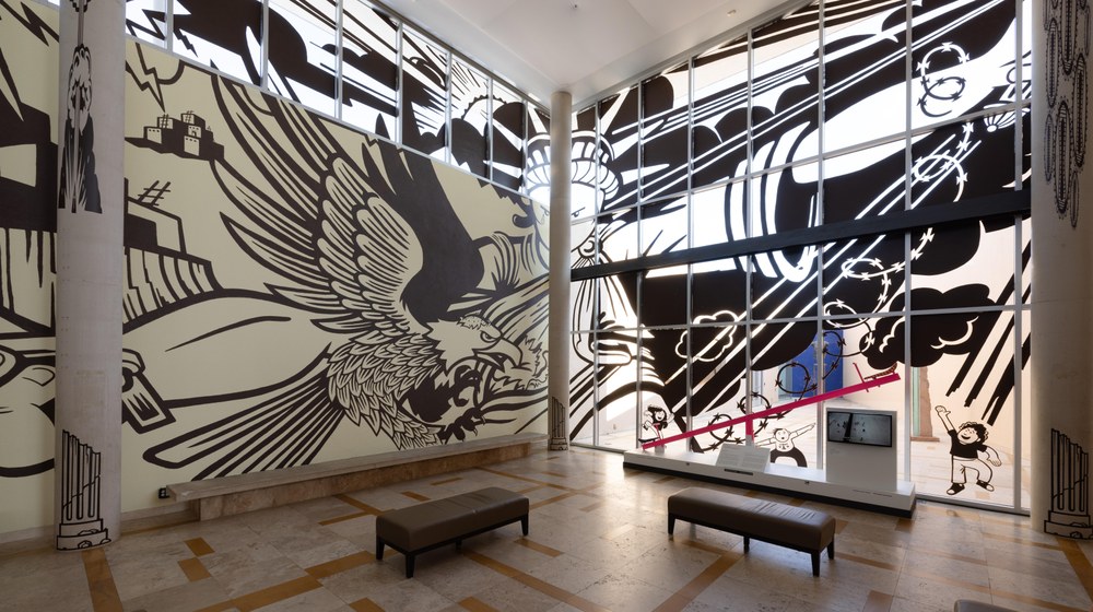 A mural on the wall and window of the Albuquerque Museum lobby. On the left is a pueblo building on a mesa. In the center is the Statue of Liberty in chains. On the right are two children running towards Teeter-Totter Wall (2019) by Ronald Rael, Virginia San Fratello, and Colectivo Chopeke.