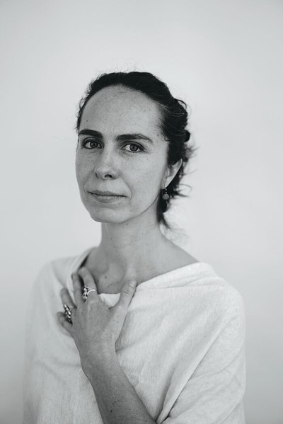 A black-and-white headshot of artist Nicola López. She has light-colored skin and dark hair, worn up. She wears a white T-shirt and rings and stands against a white background with her hand on her chest.