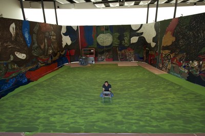 Gronk with set piece for Griselda A photograph of the artist Gronk, sitting in the middle of a large, non-objective mural across three walls with a green floor.