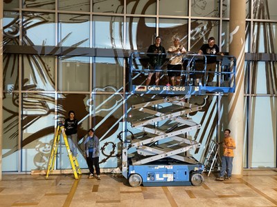 Since 1776... (in progress) A group of six people standing on the floor and on a scissor-lift in front of a projected image of Eric J. García's mural. The mural is in black and white and projected on glass. The people are smiling and wearing paint-covered clothes.