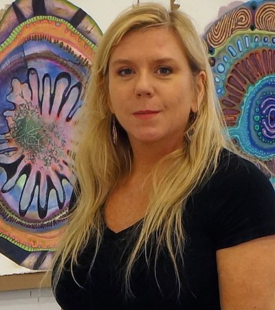 A headshot of artist Lea Anderson, who has long, blonde hair, wears a black T-shirt, and stands in front of amoeba-like artworks.