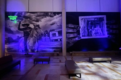 A photo of Chip Thomas' panaceas, promises + problems, taken at night. Blacklights illuminate the front of the installation, causing the small pieces of uranium hung in front of the large-scale photographs to glow green. In the top left corner of the installation, a starburst shape and text which reads "Atomic Age" also glow bright neon green.