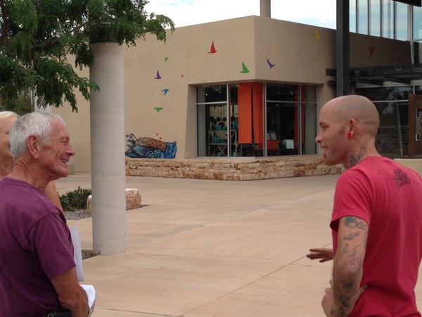 A photo of artist Ernest Doty speaking with two visitors in front of his mural on the exterior of the Albuquerque Museum. The mural features numerous small, colorful birds flying out of the body of a dead bird.