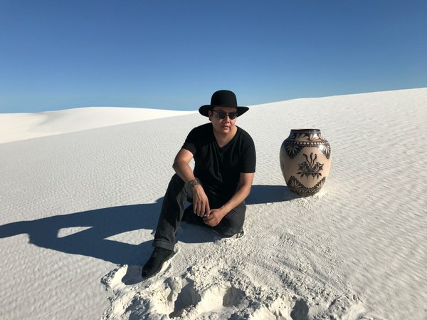 A photograph of artist Virgil Ortiz sitting on a sand dune at White Sands. He wears a wide-brimmed hat and sunglasses and sits next to a ceramic jar.