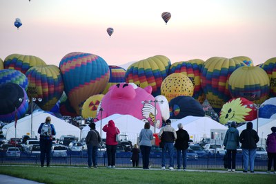 Spectators watching rows of colorful hot air balloons rising on a field.