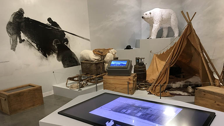 A view of the Arctic Air exhibit featuring a display of cold weather camp with a polar bear statue, a tent with furs inside, and a digital table display on a stand in the foreground.
