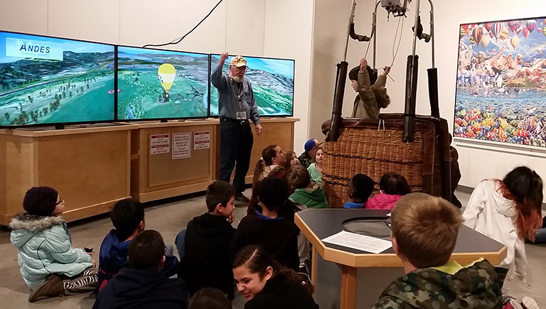 An older gentleman give a presentation to elementary-aged children who are seated on the floor in front of him. Behind him are two screens displaying aerial views of mountains and large open green fields. A small, modern hot air balloon basket is near the gentleman. A child is standing inside it while listening to the presentation.