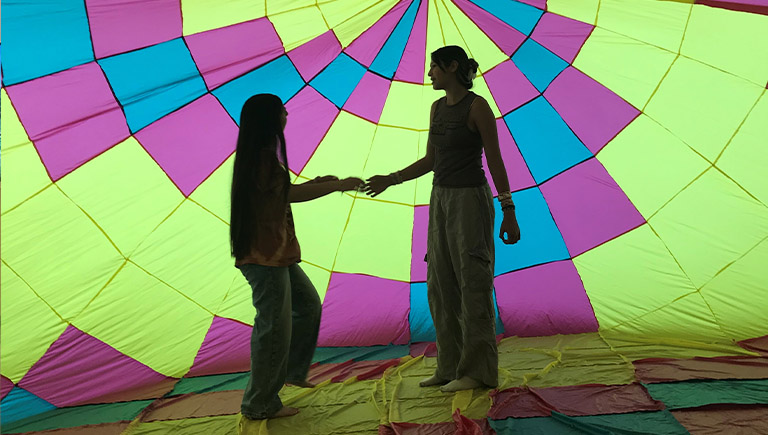 An older gentleman give a presentation to elementary-aged children who are seated on the floor in front of him. Behind him are two screens displaying aerial views of mountains and large open green fields. A small, modern hot air balloon basket is near the gentleman. A child is standing inside it while listening to the presentation.