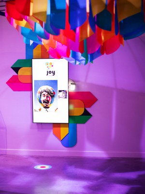 A digital display featuring an AI-generated image of a stylized face on a hot air balloon on a digital monitor, hung portrait-orientation, atop a flat, colorful artwork with geometric panels arranged vertical and horizontally.