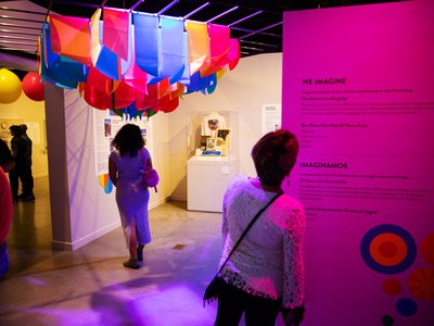 People walking through the 20 Years of Joy exhibition. One person is reading text about the exhibition printed in black text on the wall. Another is walking toward a display case with various print material and a t-shirt on a bust. Above her is a series of colorful cloth segments hung in rows.