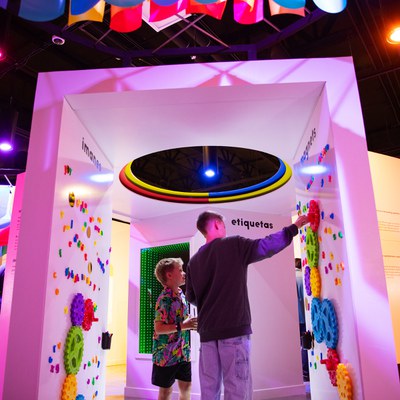 Two children interact with an exhibit in the 20 Years of Joy exhibition, which features two white walls covered in colorful plastic gears and plastic letters.