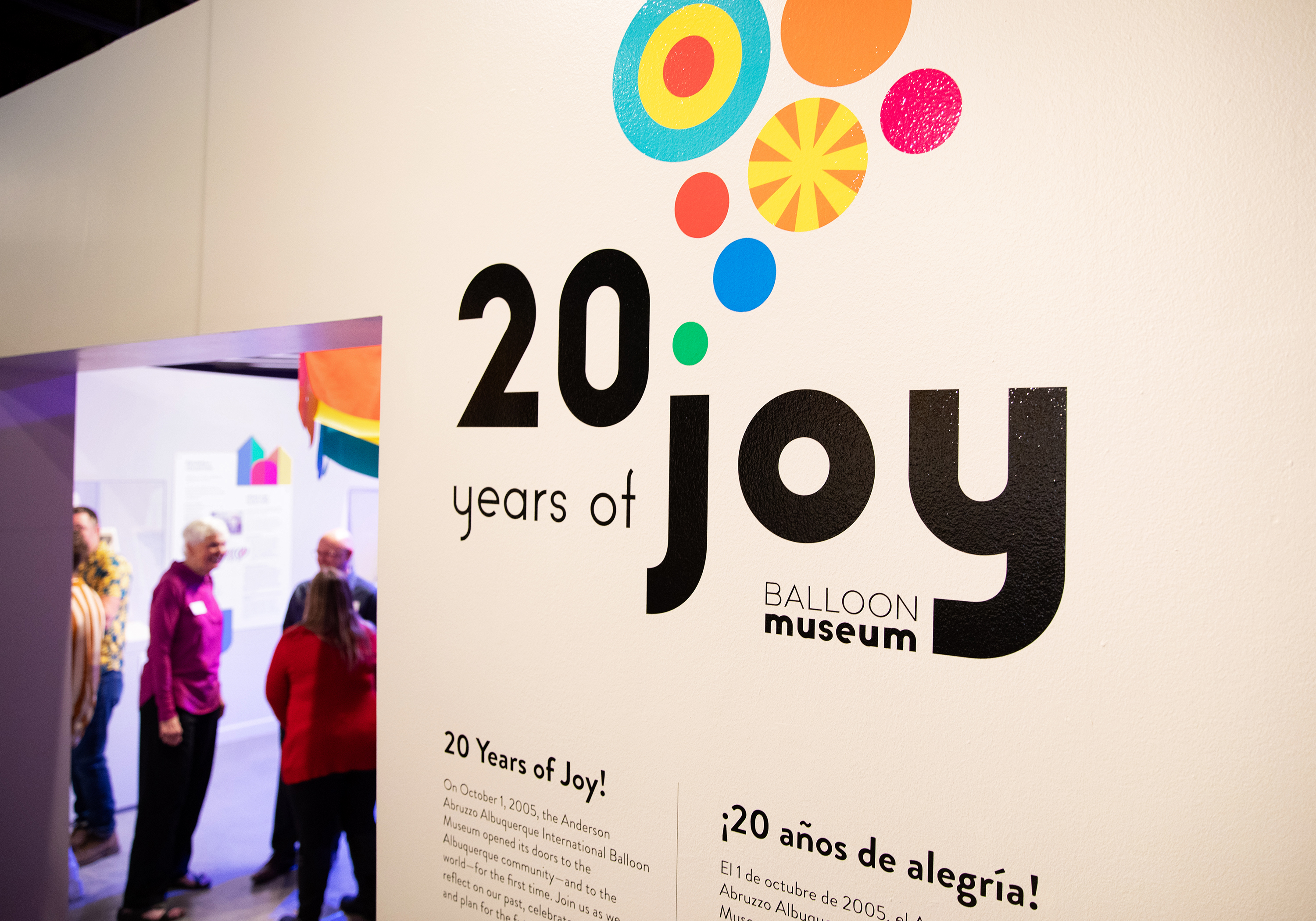 A close-up of the 20 Years of Joy Exhibition logo wall, featuring the logo above two columns of black text on a white wall that describes the exhibition. On the left-hand side is a small group of people exploring the exhibition.