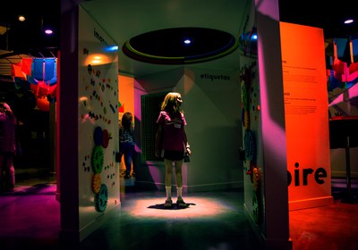 A child within an exhibit in the 20 Years of Joy exhibition, which features two white walls covered in colorful plastic gears and plastic letters. The lights have been turned down and the exhibit and surrounding area is dark, with colorful red and white spotlights aimed at the walls and floor.