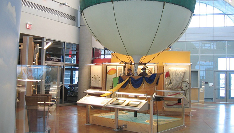 The Early Ballooning exhibition, featuring a small scale model of an early balloon with a wooden, boat-like "basket" and two men piloting the craft.