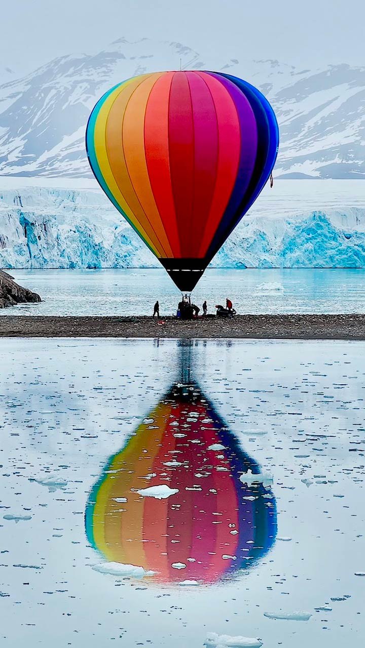 BM-Polar-Expedition(LOG-6-21-26)-web45 The Polar Expedition team with their hot air balloon fully inflated, surrounded by snowy mountains and water with small pieces of melting ice.