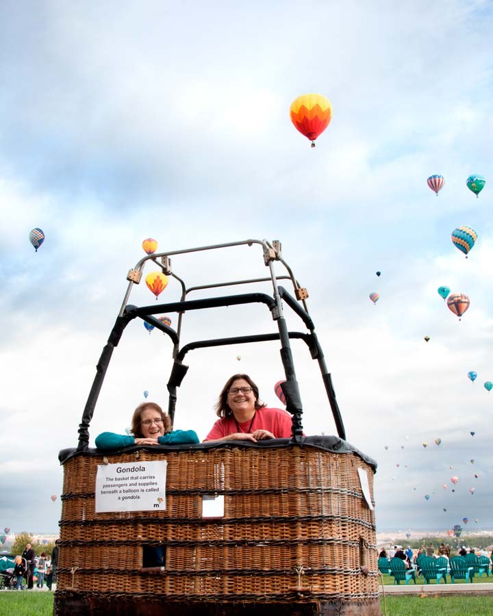 Two volunteers inside a gondola with hot air balloons floating in the sky behind them.