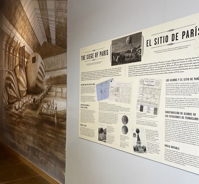 A close-up of an illustration of men working on hot air balloons during the 20th century. Next to it is a faux 20th century newspaper informational display about the exhibit.