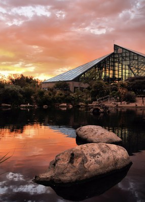 The ABQ BioPark conservatory at the Botanic Garden is pictured at sunset with a pond and two large rocks in the foreground reflecting the vibrant pink and orange light of the sunset.