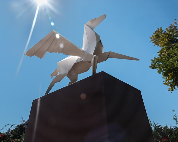 A photo looking up at large metal sculpture made to look like an origami pegasus sits on top of a large, dark base with the shining sun and blue sky above it