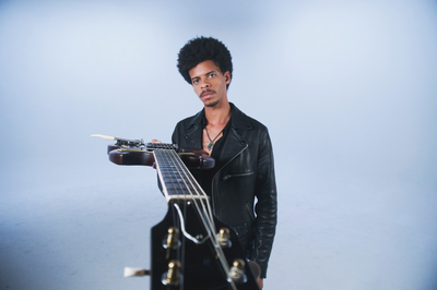 Photo of man standing in front of light blue background. He is holding out his guitar with the neck reaching out to the camera.