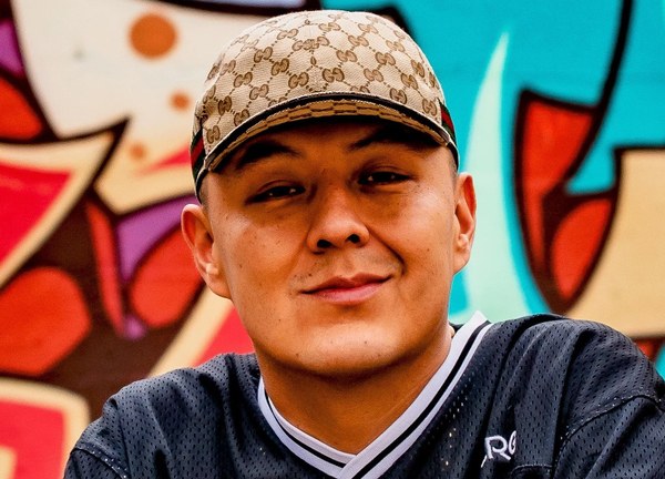 Def-i Close-up of a man with a patterned Gucci hat and dark athletic shirt standing against a vibrant urban mural.