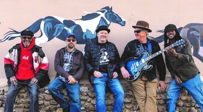 Photo of five men leaning against an adobe wall, looking toward the camera. There is a horse mural painted on the wall behind them.