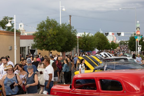 Crowd of people walking and mingling along a street lined with classic cars on display under cloudy skies.