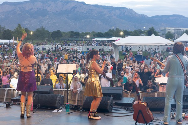 ABBAquerque performs on an outdoor stage with a large, attentive crowd and mountain backdrop at dusk.