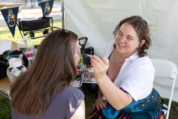 A woman with curly hair painting colorful designs on a girl's face at an outdoor event booth.