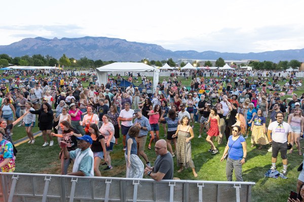 Large crowd enjoying an outdoor event on a grassy field with mountains in the background under a partly cloudy sky.