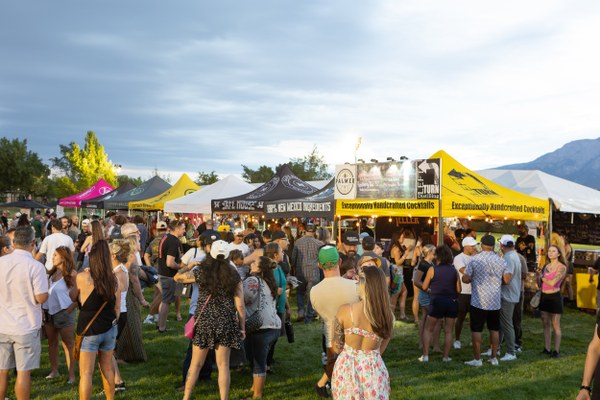 Crowd gathered at an outdoor festival with colorful tents offering handcrafted cocktails and food under a cloudy sky.