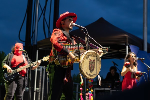 Doctor Nativo in a red hat and jacket playing guitar and singing on stage during an outdoor concert at dusk.