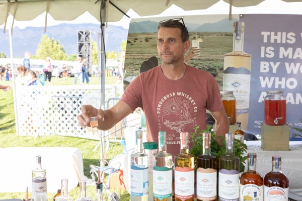 Man in a gray cap sampling a drink from a vendor at an outdoor festival booth with various liquor bottles displayed.